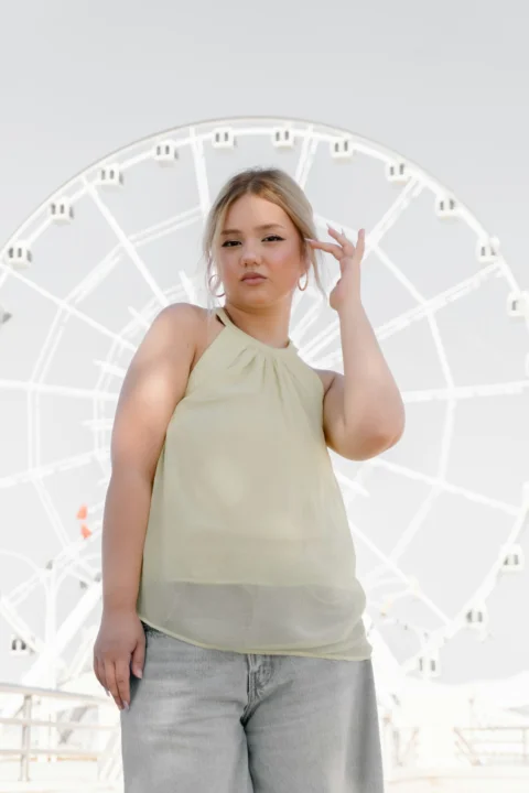 A woman in a light yellow halter top and grey jeans before a ferris wheel