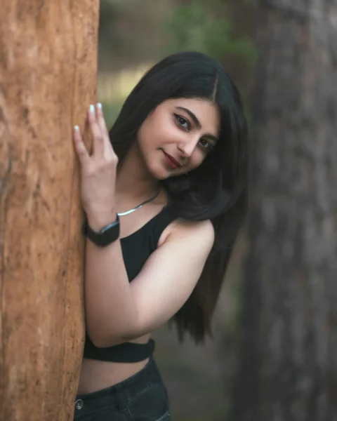 A woman in a black top leaning against a tree trunk in a forest