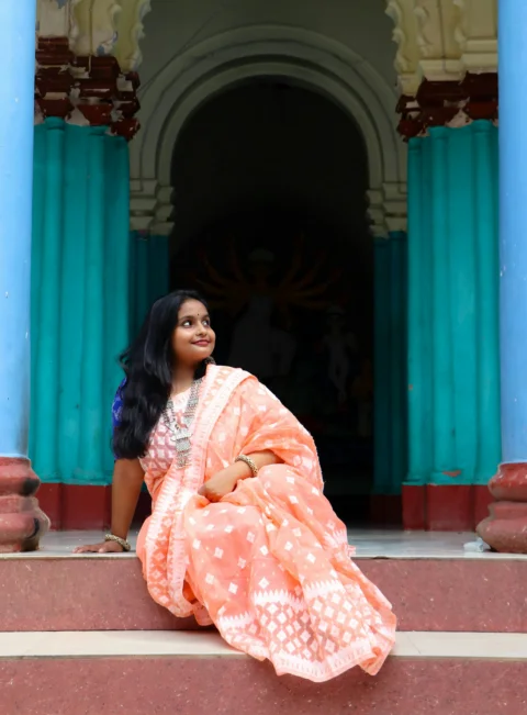 A woman in a peach sari sitting between teal pillars at a temple entrance