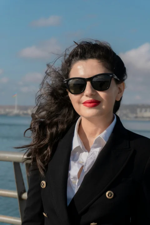 A woman with dark hair wearing sunglasses and a black blazer by the sea
