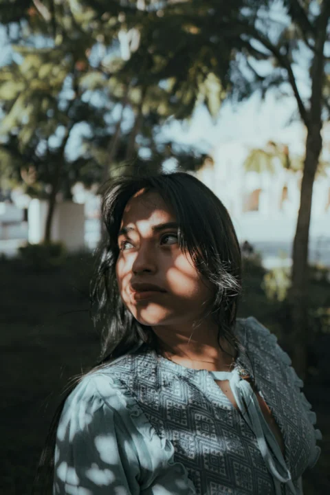 A woman poses outdoors, shadowed by trees