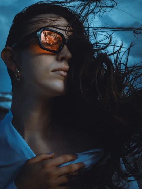 A woman with windblown hair wearing sunglasses reflecting a warm sky at night