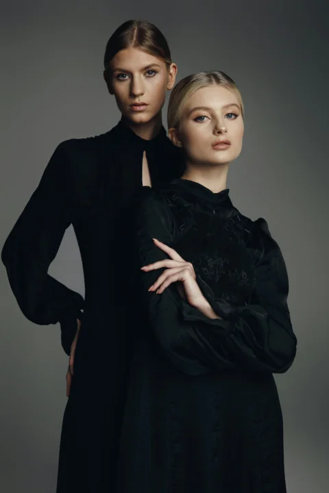 Two women in black dresses posing against a grey background