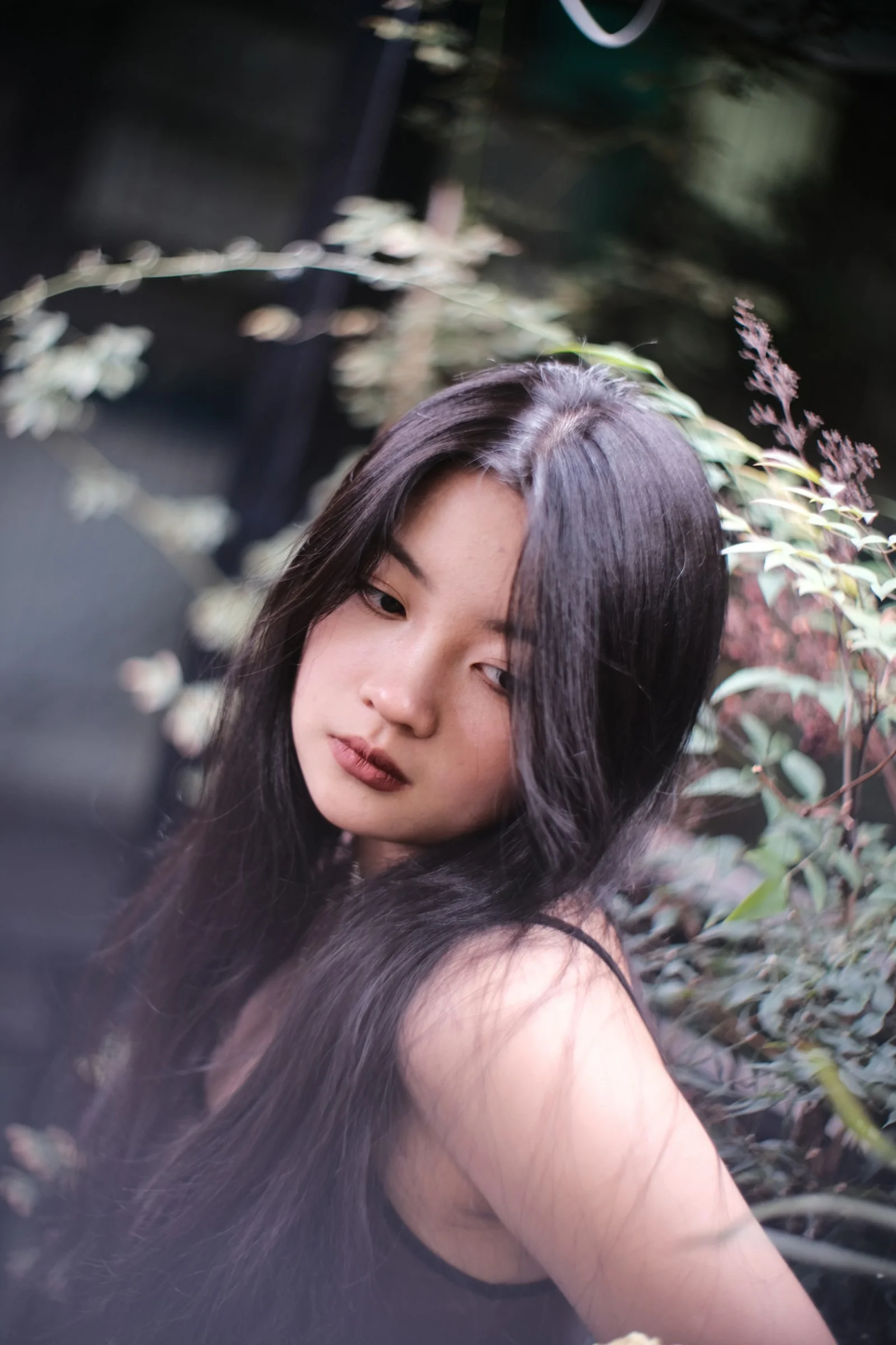 a woman with long black hair standing next to a bush