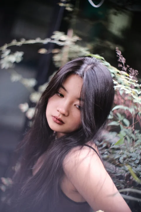 a woman with long black hair standing next to a bush