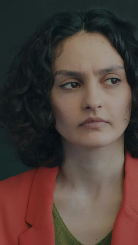 A woman with dark curly hair wearing a coral blazer and green top