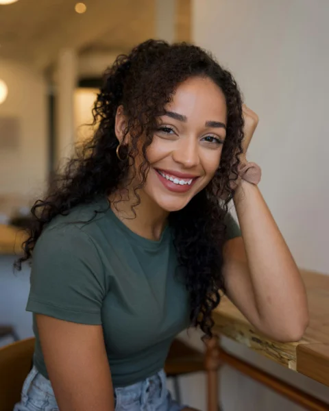 A smiling woman with curly hair wearing a green t-shirt at a wooden table