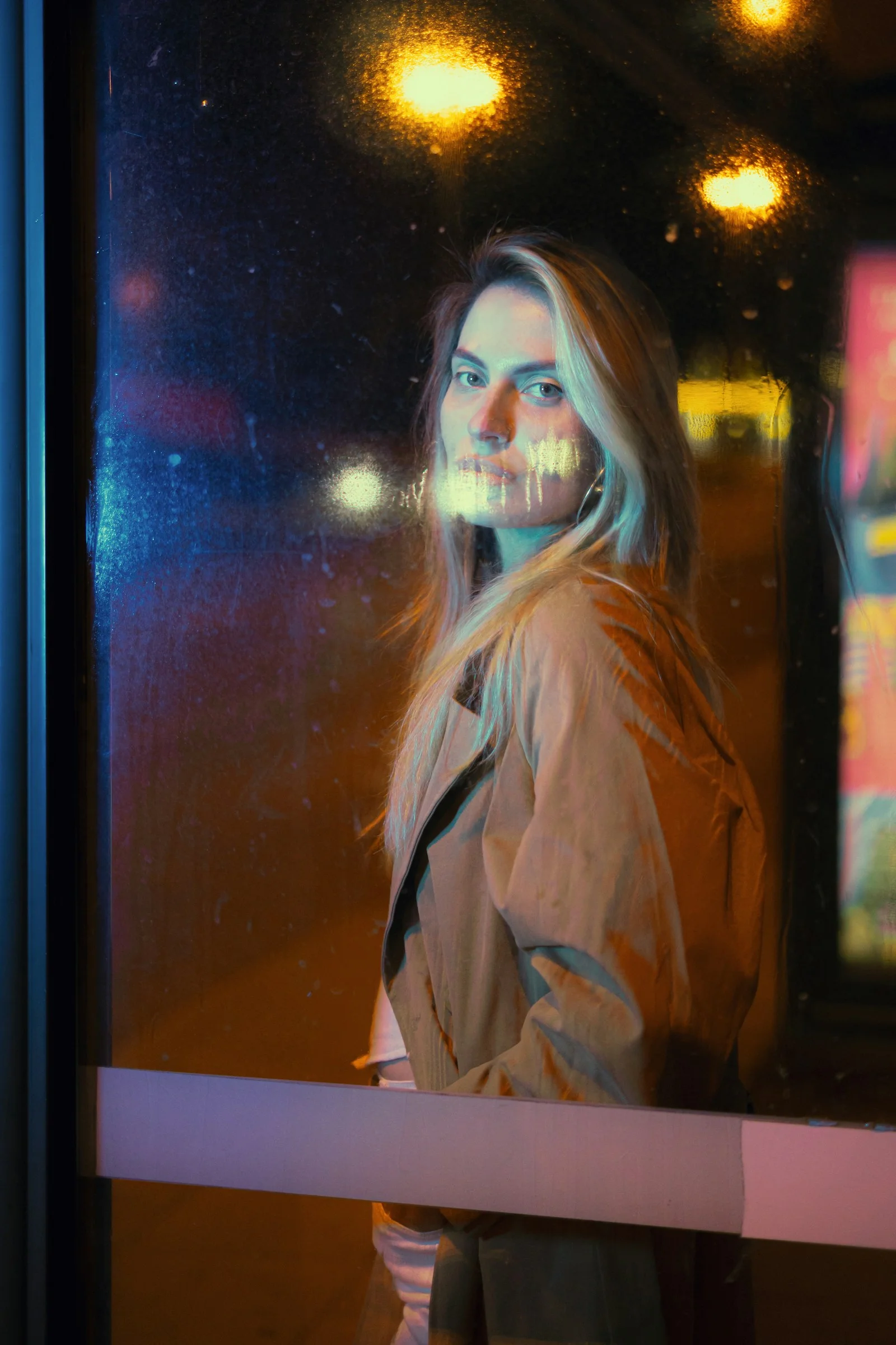 A woman in a tan coat behind frosted glass with blue and orange light