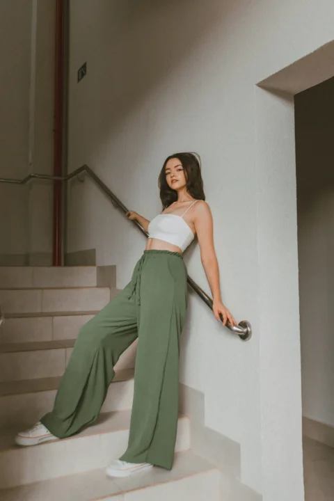 a woman in a white top and green pants leaning on a stair rail