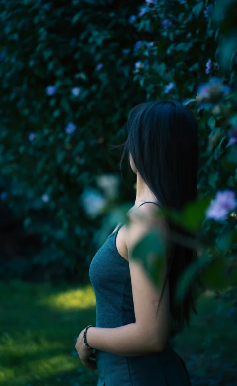 グリーンのタンクトップを着た長い黒髪の女性が花の咲く茂みのそばにいる