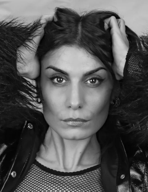 A woman with hands in her dark hair looks forward