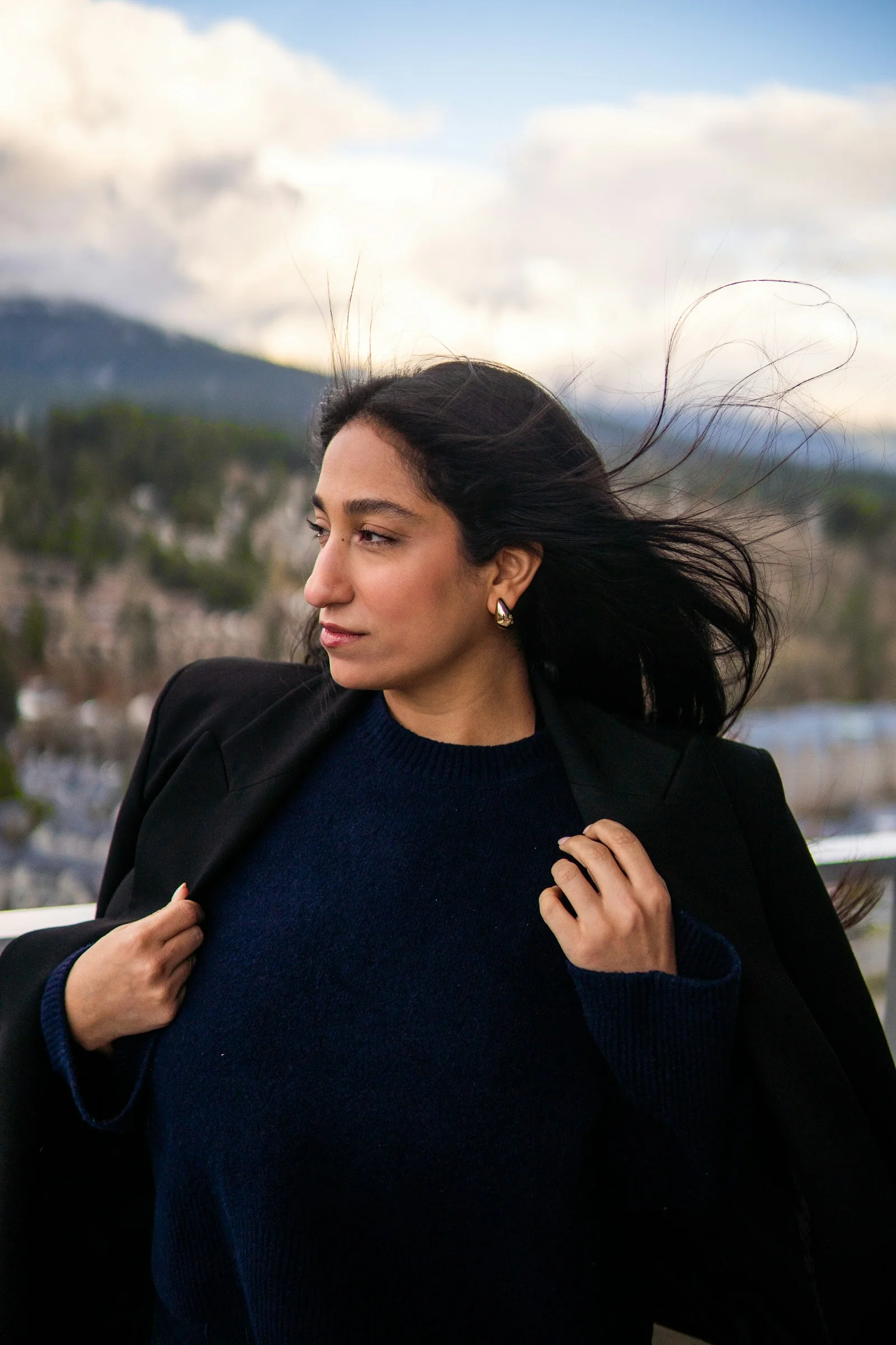 A woman in a dark sweater and blazer with windblown hair outdoors