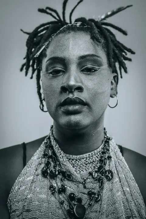A person with dreadlocks and necklaces gazes directly forward