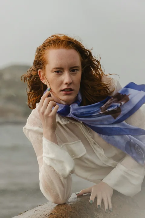 A woman with red hair wearing a white shirt and blue scarf outdoors