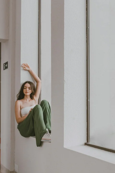 a woman in a white top and green pants sitting on a window sill