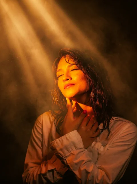 A woman with closed eyes in a white shirt illuminated by a diagonal beam of warm golden light