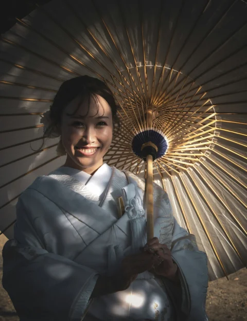 A smiling woman in a white kimono holding a traditional paper umbrella in warm light