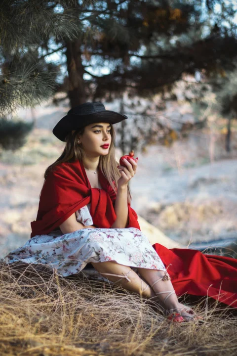 a woman in a red dress and black hat is sitting on the ground