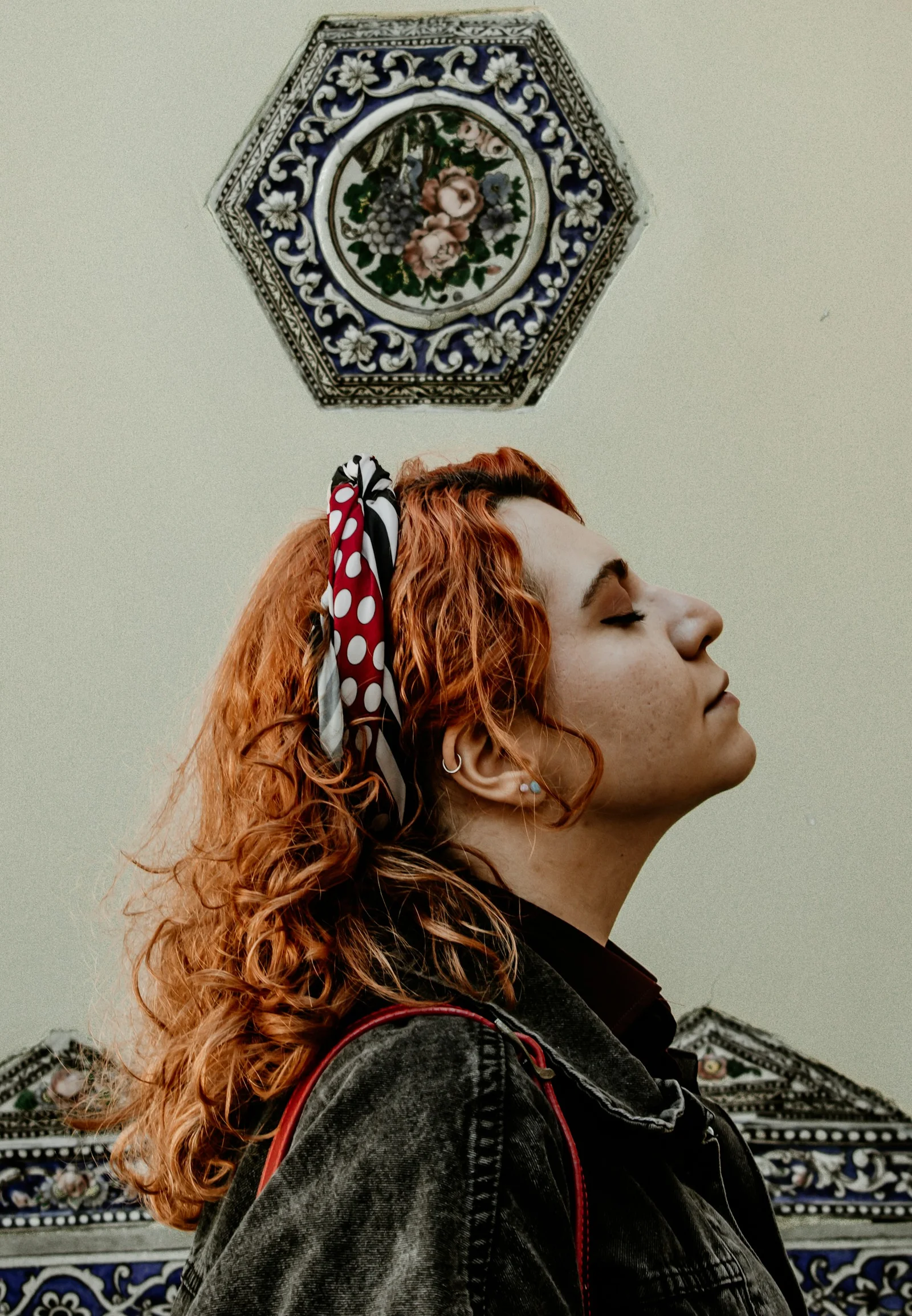 A woman with curly red hair and a polka dot headscarf against a tiled wall