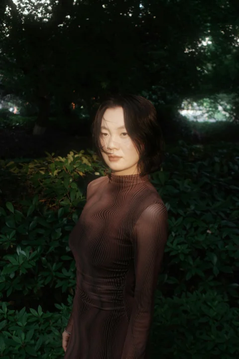 A woman in a brown patterned long-sleeved top standing before dark green foliage