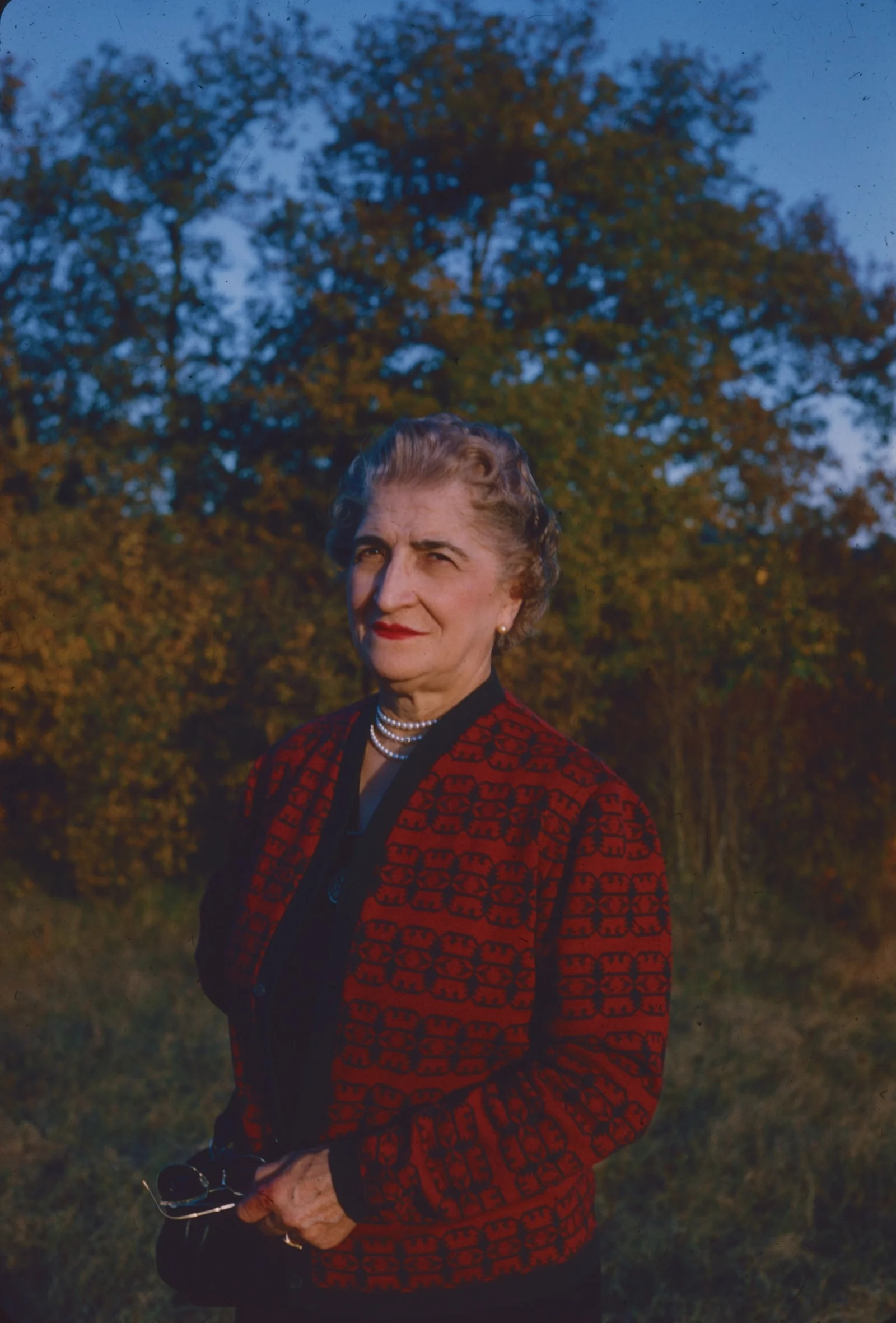 An adult woman in a red patterned jacket and pearl necklace outdoors at golden hour