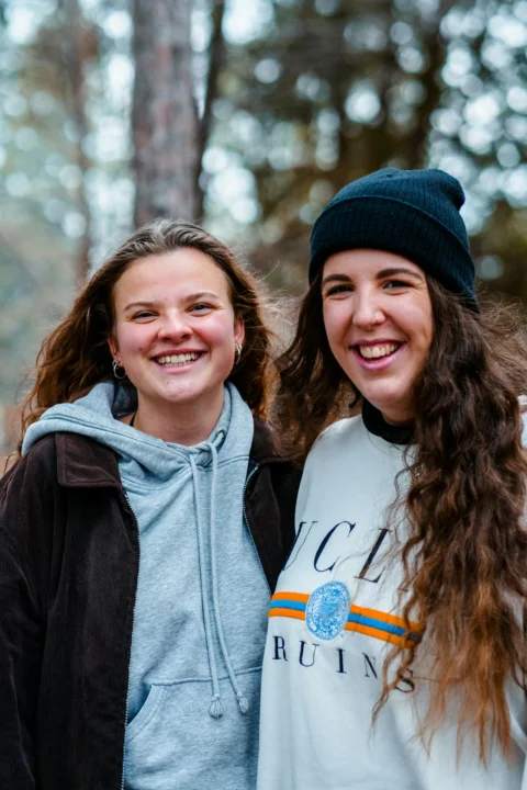 Two women smiling in a forest, one wearing a grey hoodie and brown jacket