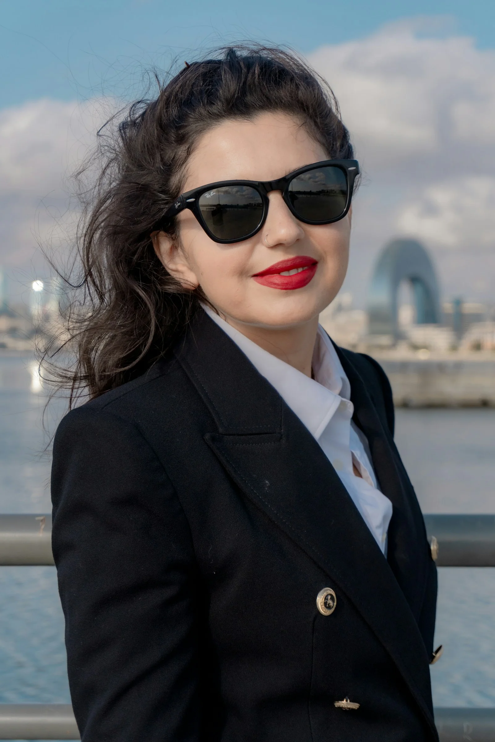 A woman in a black blazer and white shirt wearing sunglasses by the water