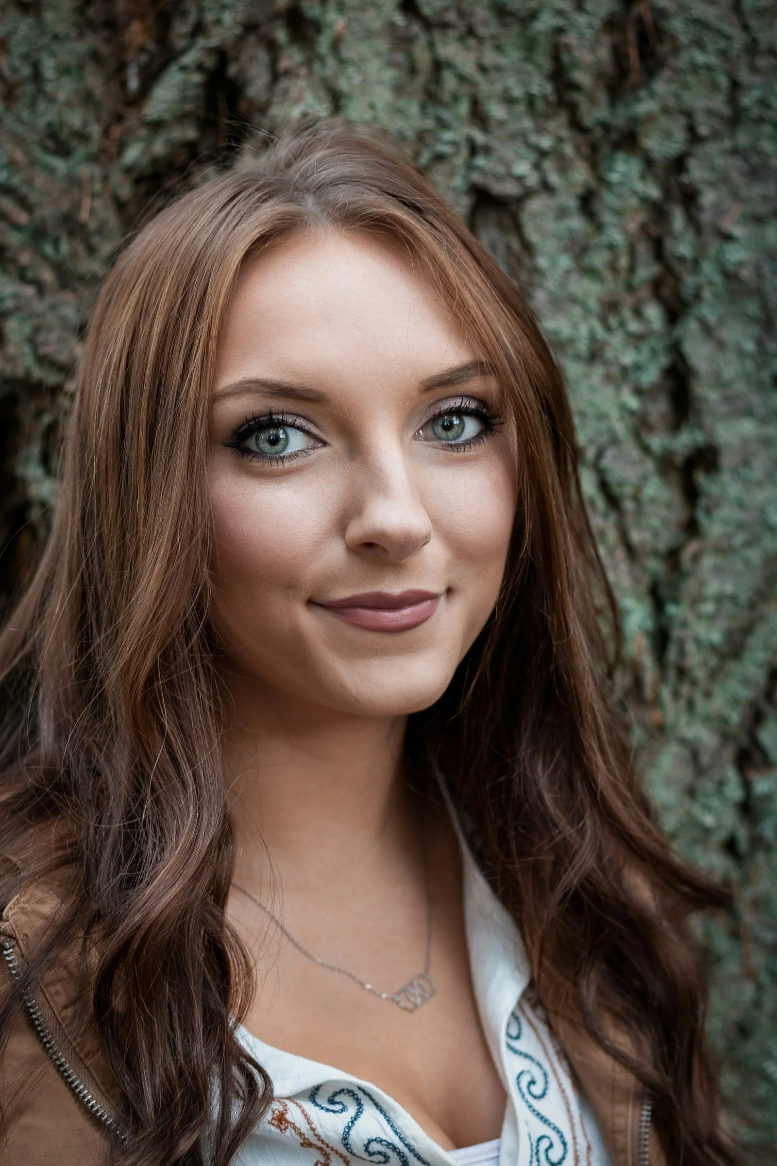 a woman with long hair and blue eyes posing in front of a tree
